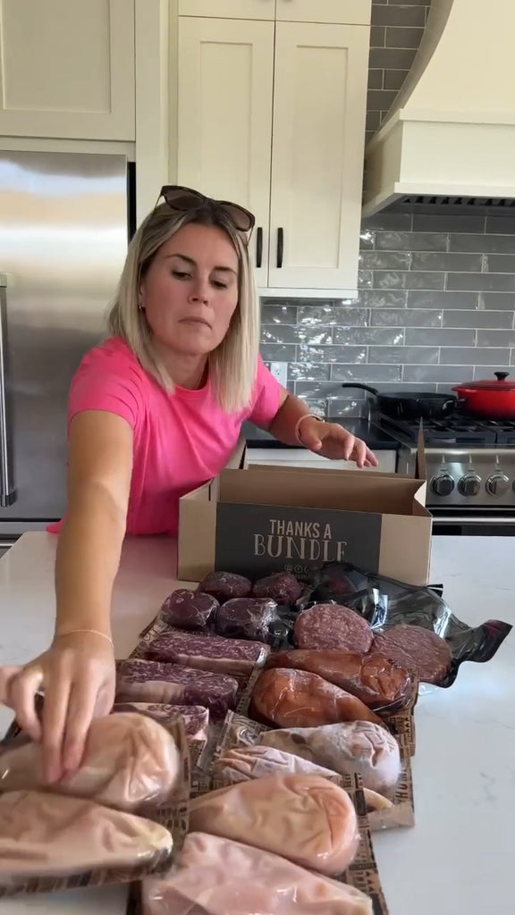 A blonde woman in a pink shirt unpackaging meats from a Butcher Bundle and laying them out neatly on a kitchen island. Items from the meat box include grass-fed and finished steaks, and free run small batch marinaded chicken breasts.