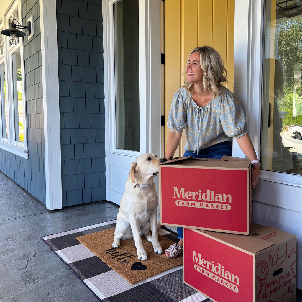 A blonde woman with her golden retriever happily accepting two red Meridian grocery delivery boxes on her front porch.