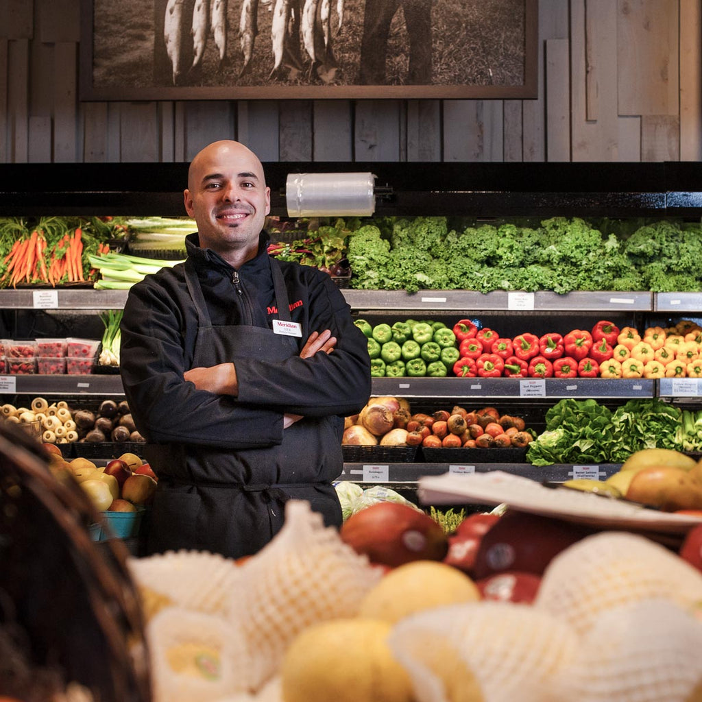 A male produce manager wearing a black apron standing in front of fresh, farm market fruits and vegetables.